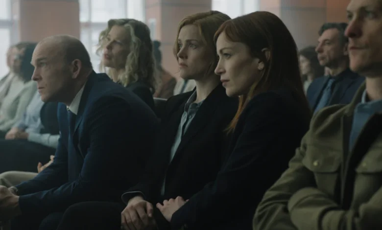 "People sit in rows in a courtroom or formal setting, dressed in business attire, attentively focused on proceedings taking place out of frame, with large windows and neutral lighting in the background."